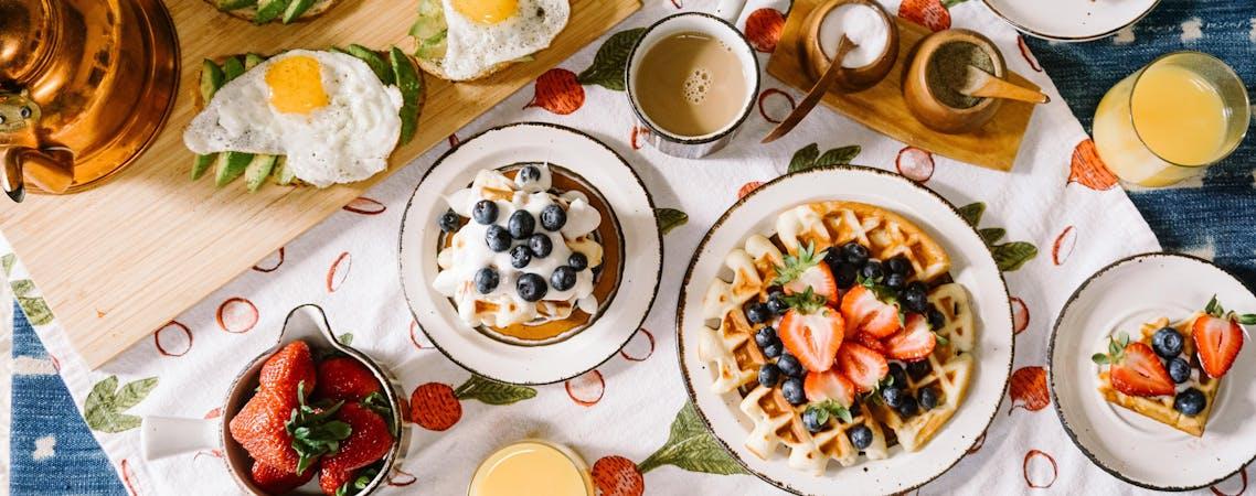Foto de mesa com pratos redondos de cerâmica branco cheio de waffle, frutas, café e suco.