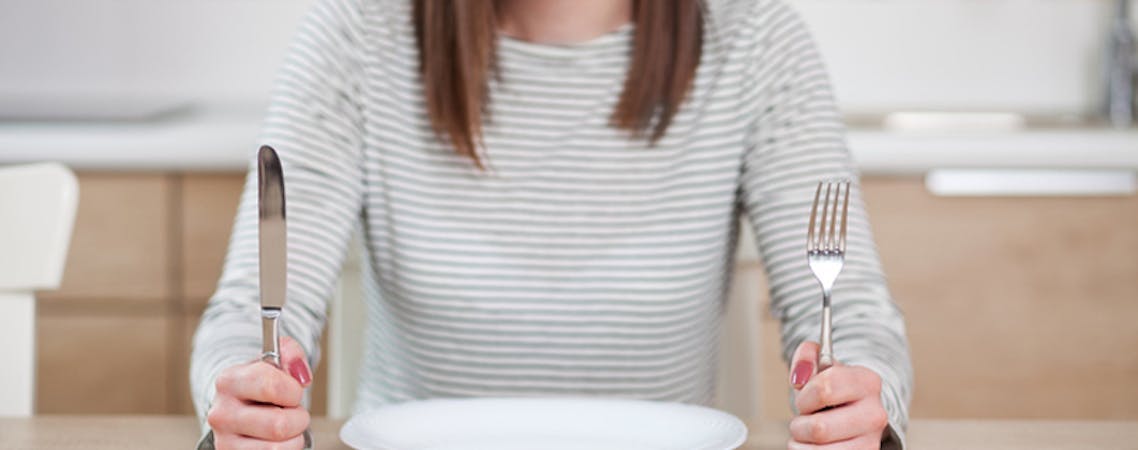Foto de mulher sentada em uma mesa com um prato de comida vazio segurando o garfo e faca.