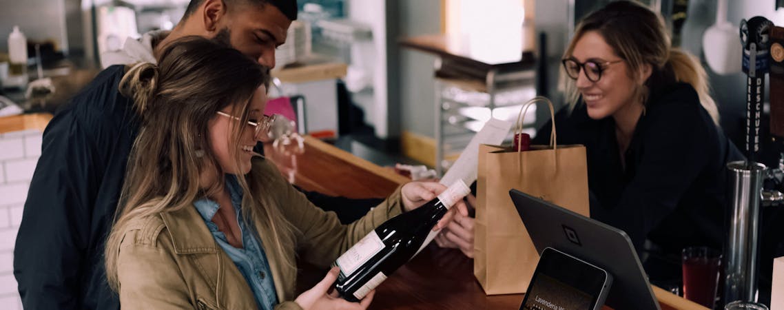 Foto mulher segurando garrafa de vinho ao lado de homem na frente de mulher sorrindo.