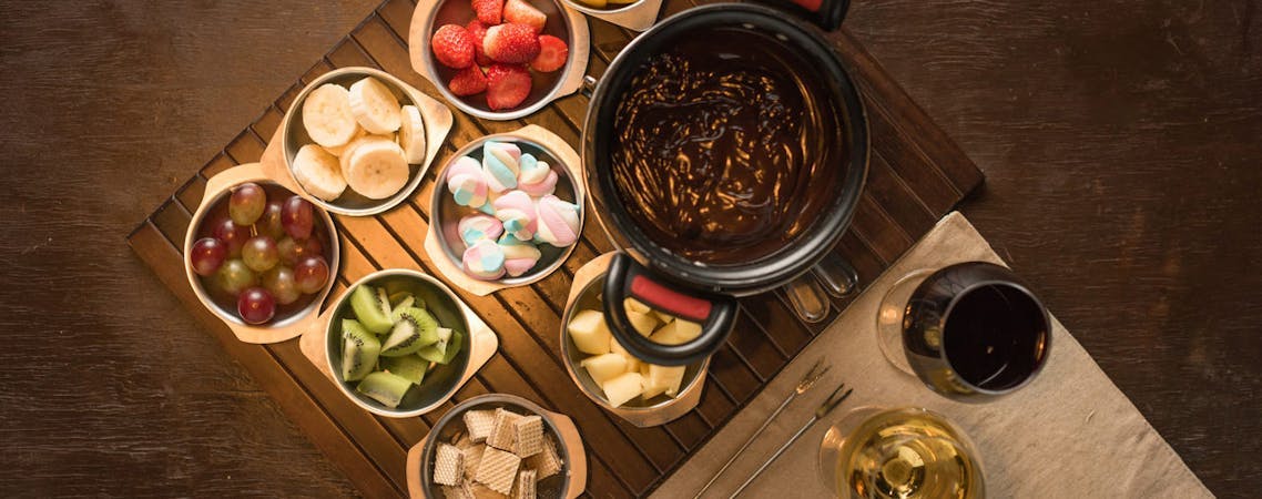 Foto de uma mesa com fondue de chocolate, frutas, e vinho.